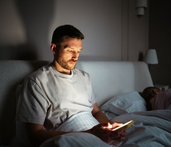 Man awake in bed at night looking at his phone while his partner sleeps beside him, illustrating disrupted sleep in the Sleep Apnoea section.